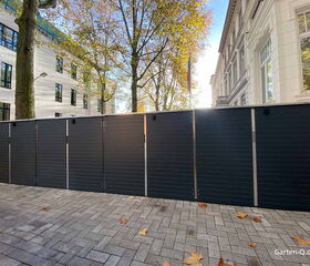 Schwarze Gartenschränke auf einem gepflasterten Gehweg, umgeben von Herbstbäumen und weißen Gebäuden.