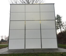 Ein modernes, weißes Gebäude mit quadratischen Paneelen, steht auf gepflastertem Boden, umgeben von Bäumen und Gras.