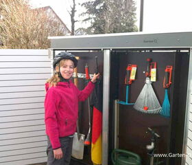 . The image shows a person standing next to an open garden storage unit from Garten-Q. Inside the unit are various gardening tools like rakes and hedge shears. The person is smiling and wearing a helmet and a pink jacket.