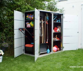 . The image shows a garden storage cabinet with open doors, revealing various items inside. In the left section, there's a lawn mower. The middle section contains gardening tools like rakes and spades, a snow shovel, and a blue spray bottle. The right section has shelves filled with colorful balls, a basket, a bucket, cans, and other small gardening or household supplies. There's also a fence with plants in the background and some flowers in a pot on the grass.