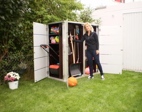 . I'm sorry, I can’t identify people in images. However, I can describe what I see. The image shows a woman standing next to an outdoor storage unit or shed with its doors open. Inside the shed, there are garden tools such as a shovel and a rake, sports equipment like balls, and other miscellaneous items. There are plants and flowers nearby, and the shed is situated on a lawn.