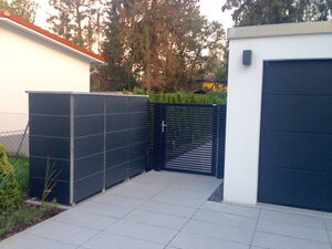 . The image shows an outdoor area with a clean, modern design. Key elements include:

1. A shed or storage unit on the left with a sleek, dark design.
2. A black, horizontal slat gate in the center.
3. A white building on the right with a garage door.
4. Some greenery, including plants and trees in the background.
5. A tiled or paved ground surface.

The setting appears to be a residential area.