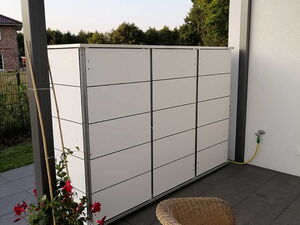 . The image shows an outdoor patio area that includes a modern storage cabinet with a grid-like design. The cabinet is situated against a wall, supported by a structure with a transparent roof. There's a potted plant with red flowers next to the cabinet, and a wicker chair is placed in front. The patio is paved with large, dark tiles, and there's a garden hose attached to the wall in the background.