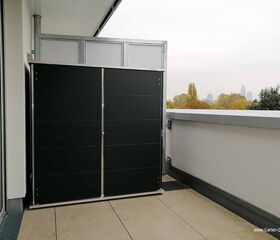 . Ein Balkon mit einem schwarzen Sichtschutz oder Schrank, Stadtansicht im Hintergrund.
