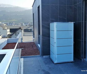 . Ein moderner Balkon mit einem weißen Schrank, Gebäuden im Hintergrund und Bergen in der Ferne.
