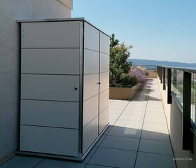 . Ein weißer Gartenschrank steht auf einer Terrasse neben einem Geländer, mit Pflanzen im Hintergrund.