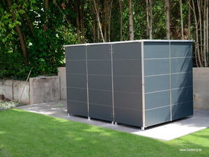 . The image shows a modern, dark-colored garden shed or storage unit situated in a backyard. It is placed on a paved area surrounded by grass, with trees and shrubs visible in the background. The shed has a sleek, minimal design with metal or composite panels.