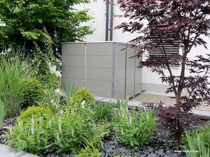 . The image shows a garden scene with a modern storage box or shed placed against a white building. There are various plants and shrubs in the foreground, including both green and purple foliage. The ground is covered with small dark rocks or gravel, adding to the landscaped appearance.
