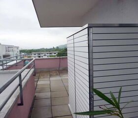 Eine überdachte Terrasse mit Metallgeländer, Fliesenboden und angrenzenden Gebäuden bei bewölktem Himmel.