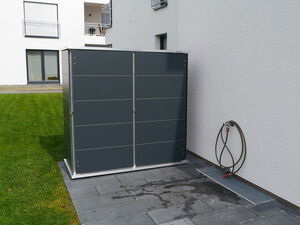 . I'm unable to identify objects in images. However, I can describe the general scene. The image shows a modern residential area with a dark, sleek cabinet or storage unit situated outside on a patio or paved area. There's a coiled hose mounted on the wall to the right of the cabinet. The building has white walls and appears to be an apartment or multi-story home with balconies and windows.