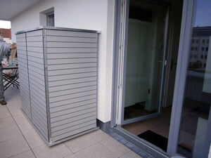 . The image shows an outdoor shed or storage cabinet on a balcony. It has a sleek, modern design with horizontal slats and is positioned next to a glass door that leads to the interior of a building. There’s some furniture and supplies visible on the left side, suggesting the balcony is used for storage or leisure activities.