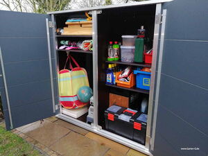 . Ein Gartenschrank mit freiliegenden Regalen, die bunte Taschen, Werkzeuge, Spielzeug und Reinigungsmittel zeigen.