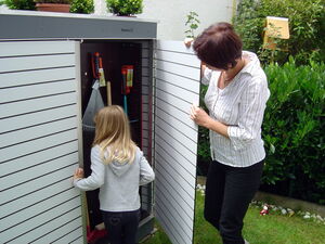 . The image shows two people, an adult and a child, opening a garden storage shed. The shed contains tools like a rake and a saw. It's set in a yard with plants and a lawn visible around it.
