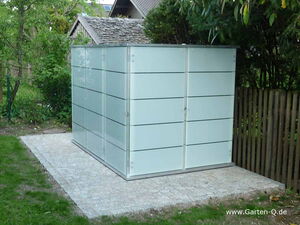 . The image shows a modern, cube-shaped garden shed or storage unit with a sleek design. The walls appear to be made of frosted glass or a similar material with horizontal lines. It's set on a paved area in a garden, surrounded by grass, trees, and a wooden fence.