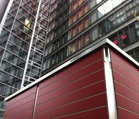 . The image shows a modern architectural scene with a focus on a red panel structure in the foreground. Behind it, there is a tall, glass-fronted building with multiple windows. The urban setting suggests a city environment, and the structure has a metal frame. The words "Garten Q" are visible on the structure.