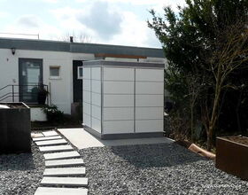 . The image shows a modern, minimalist garden setting. In the center, there is a sleek white storage shed with horizontal paneling. A pathway made of rectangular slabs leads to this structure, surrounded by gravel. The area is landscaped with some greenery and a planter box is visible to the left. In the background, part of a house with a small window and door can be seen.