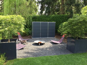 . The image shows an outdoor area with a setting that includes the following elements:

1. Two modern lounge chairs with colorful patterns.
2. A metal fire pit at the center of the chairs.
3. Two large square planters with small trees.
4. A gravel surface covering the area.
5. A dark-colored structure, possibly a shed or storage unit, in the background.
6. Lush greenery and trees surrounding the area, providing a natural backdrop.
7. A cat standing near one of the lounge chairs.

This arrangement appears to be a well-designed outdoor relaxation or lounge space.
