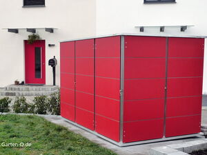 . The image shows a modern outdoor setting. There's a large red modular structure, possibly a garden storage unit or shed, on a paved area. In the background, there's a building with a red door, a mailbox, and some decorative plants. The ground in front is landscaped with grass and shrubs.