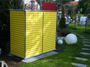 . Ein gelbes Gartenhäuschen aus Metallplatten steht in einem grünen Garten mit Pflastersteinen und Kugeldeko.