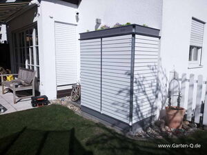 . The image shows a garden scene with a modern storage unit against the exterior of a white house. The unit is sleek with horizontal slats, resembling a garden tool shed. There's a patio area with a wooden bench and various tools or equipment around. Potted plants and a small section of a white picket fence can also be seen.
