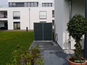 . The image depicts a modern outdoor setting. Elements include:

1. A white multi-story building in the background with balconies and windows.
2. A large green lawn.
3. A paved area with gray tiles.
4. A small tree or shrub in a pot on the right.
5. A dark panel or structure near the wall.
6. Outdoor furniture such as a table and chair.
7. A coiled hose or cable on the wall.
