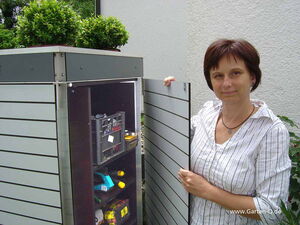 . The image shows a person standing next to an outdoor storage cabinet. The cabinet is open, revealing shelves that contain various items such as a crate, some tools, and a few other objects. On top of the cabinet, there are two potted plants. The setting appears to be outdoors, possibly in a garden or backyard area.