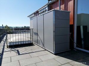 . The image shows a rooftop area with a large rectangular object, possibly a storage unit or enclosure, to the right. It is next to a railing around a skylight. The background features a view of trees and buildings. The image has a watermark displaying www.Garten-Q.de.