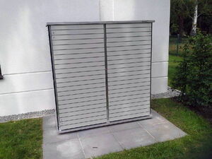 . This image shows a structure that appears to be a metal storage box or cabinet placed outdoors. It has a slatted front with two doors and is situated on a paved area next to a building, surrounded by grass and some shrubs.