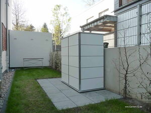 . The image shows a small garden area with a modern, rectangular storage shed. The shed is situated on a tiled section surrounded by grass. To the left, there is a small bench or table and some pebble landscaping along the edge of the building. There is fencing and some sparse shrubbery along the right side. The area is enclosed by building walls and fences.