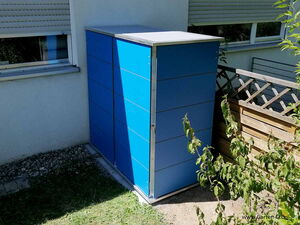 . The image shows a blue outdoor storage cabinet placed against a house wall. It's situated next to a window and partially surrounded by a wooden fence. There's a small plant or bush in front of the cabinet. The ground is a mix of grass and some stones near the wall.