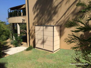 . The image shows an outdoor scene featuring a modern building with a rounded balcony and a set of stairs. There is a rectangular structure or cabinet against the wall, and the area is surrounded by greenery, including various plants and trees. The grass and shadow on the wall suggest it is a sunny day.