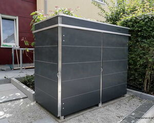 . The image shows a modern outdoor storage unit. It has a sleek design with a dark color and metallic accents. There are plants on top of it, suggesting it may also serve as a planter. The unit is situated in a garden area next to a building.
