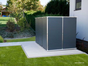 . The image shows a modern, compact metal storage unit or garden shed placed on a tiled patio in a landscaped garden. It's situated next to a building with an exterior wall. The surrounding garden area has neatly trimmed grass, shrubs, and a few trees, creating a tidy and organized outdoor space.
