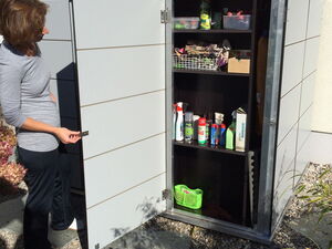 . The image shows a storage cabinet being opened, revealing various cleaning and gardening supplies. The shelves contain spray bottles, containers, and some boxes. There is also a small green basket on the bottom shelf. A person is standing next to the cabinet, opening its door.