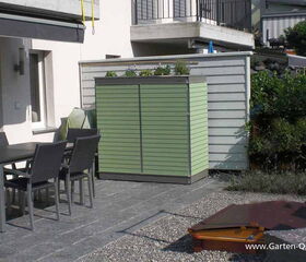 . The image shows a patio area with outdoor furniture, including a black table and chairs, situated on a tiled surface. Next to the furniture is a garden storage unit with a green facade. The area appears to be part of a backyard or outdoor space adjacent to a building, with some plants around the storage unit and nearby structures. There's also a canopy or awning above the patio.
