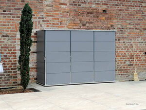 . Ein grauer Gartenschrank vor einer Ziegelwand, daneben eine schlanke Pflanze und ein Besen.