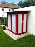 The image shows a small outdoor storage shed with bright red walls and white panels. It has multiple doors with handles and is situated on a tiled area, surrounded by grass. There's a small potted plant nearby, and the shed is adjacent to a wall in a residential area with some houses and trees visible in the background.