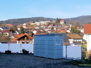 . Eine Dachterrasse mit Pflanzenkästen und einem weißen Schrank im Vordergrund, Stadtlandschaft im Hintergrund.