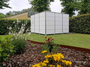 . Ein moderner Geräteschuppen aus weißen Platten steht in einem Garten mit bunten Blumen und grüner Hecke.