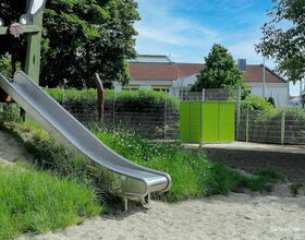 Eine Rutsche auf einem Spielplatz, umgeben von Gras und Bäumen, mit einem Zaun und Gebäude im Hintergrund.