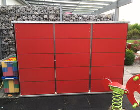 . The image shows a red storage unit or cabinet with metallic borders and handles. Behind it is a wall of rocks contained in a metal mesh. To the side, there is a stack of large colorful building blocks in a transparent container. In the foreground, there is a red and green playground spring rocker. The scene appears to be in an outdoor area with some greenery visible in the background.