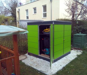 . The image shows a small green garden shed in a backyard. The shed door is open, revealing garden tools and equipment inside, including a hose and what appears to be a lawnmower. The shed is placed on a gravel area surrounded by grass. Nearby, there are various garden structures, a compost bin, and a building in the background.