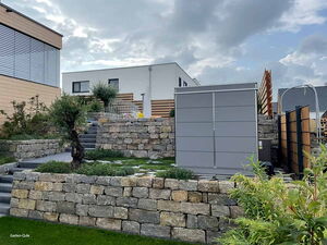 . Terrassierte Gartenlandschaft mit Natursteinmauern, grauem Schuppen, Treppe, Pflanzen und modernem Haus im Hintergrund.