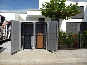 . The image shows an outdoor setting with two trash bins housed in a metal enclosure. The enclosure has doors open, allowing access to the bins, one is black and the other is brown. There's a tree nearby, and part of a building and vehicle can also be seen in the background and foreground.