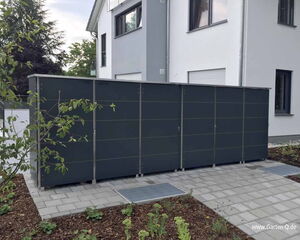 The image shows a large, dark-colored outdoor storage unit or shed next to a modern house. The structure is positioned on a paved area, surrounded by landscaped garden beds with mulch and plants. The background includes a white and gray house with windows.