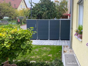 Ein Paketkasten kombiniert mit Mülltonnenboxen im Vorgarten neben einem Haus mit Blumen auf der Fensterbank und umgeben von Bäumen und Rasen.