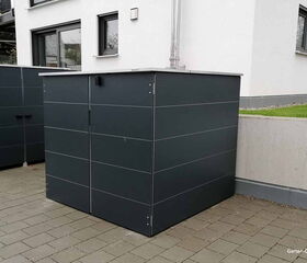 Schwarzer Mülltonnenbox vor einem modernen Gebäude, Pflastersteine ​​darunter.
