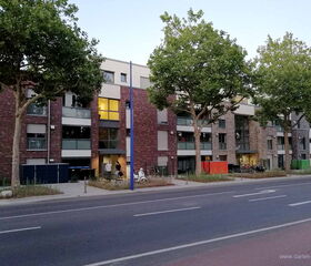 Mehrfamilienhäuser mit Bäumen und Straße im Vordergrund, einige Menschen und Fahrräder auf dem Gehweg.