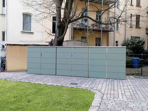 . Ein moderner Fahrradunterstand aus grauen Paneelen, im Hof mit Baum und Rasen.
