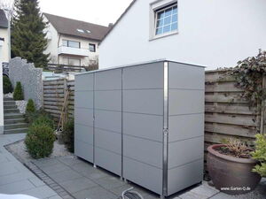 . The image appears to depict a modern outdoor storage unit or shed in a residential garden setting. It's surrounded by paving stones, plants, and a wooden fence. The unit is sleek with a gray design, suitable for storing garden tools or equipment. There are houses and some greenery visible in the background.